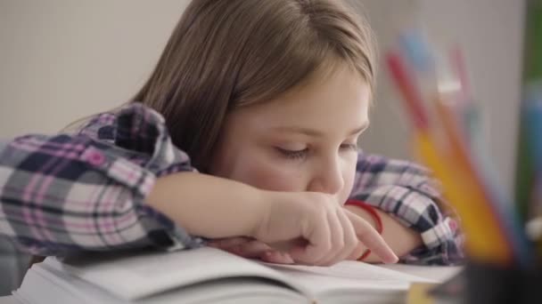 Portrait of cute brunette Caucasian girl reading book at the table. Close-up of schoolchild doing homework at home. Education, intelligence, studying. - Materiał filmowy, wideo