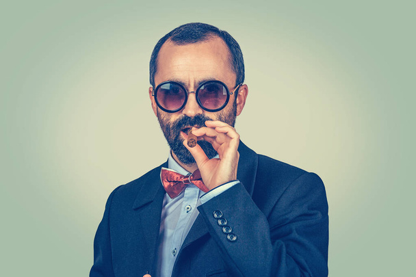 Portrait of confident, rich, bearded man with round sunglasses, keeping with hand mouth smoking cuban cigar. Arrogant boss looking to you camera. Modern clothes, dark suit, blue shirt, red bow, tie. - Valokuva, kuva