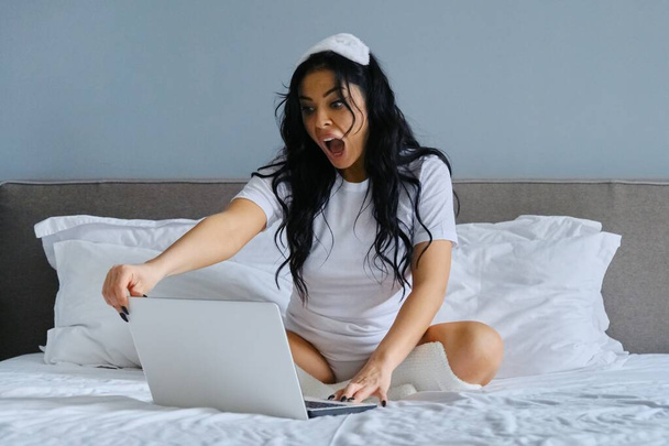 Beautiful emotional woman sitting in bed using laptop computer - Foto, afbeelding