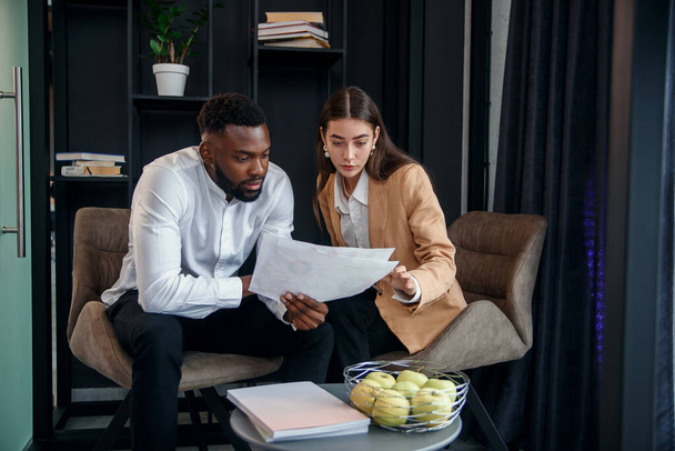 Business couple of african american man and european woman which sitting in specially designated room and using paper documents and tablet pc, conversing about their business project and their - Foto, immagini