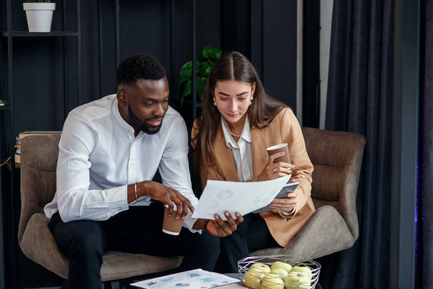 Business couple of african american man and european woman which sitting in specially designated room and using paper documents and tablet pc, conversing about their business project and their - Φωτογραφία, εικόνα