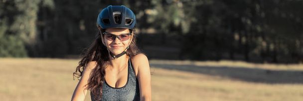 Dağ bisikletli bir kız gün batımında bir bisikletçinin güzel portresi spor kıyafetlerinde modern bir karbon fiber dağ bisikleti sürüyor. Kasklı ve gözlüklü bir kızın yakın plan portresi. - Fotoğraf, Görsel