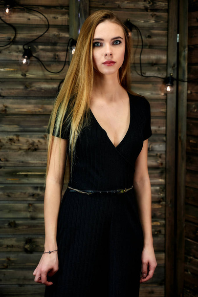 The concept of glamor, style. Vertical portrait of a pretty blonde girl with long hair and excellent make-up on a wooden background with lights. Standing in different poses in front of the camera. - Foto, Imagen