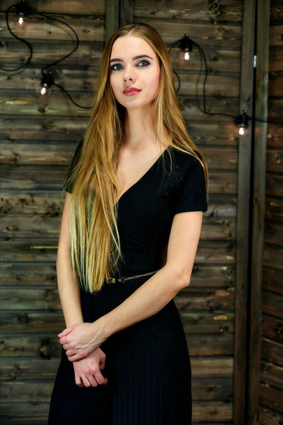 The concept of glamor, style. Vertical portrait of a pretty blonde girl with long hair and excellent make-up on a wooden background with lights. Standing in different poses in front of the camera. - Fotografie, Obrázek