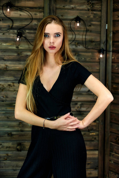 The concept of glamor, style. Vertical portrait of a pretty blonde girl with long hair and excellent make-up on a wooden background with lights. Standing in different poses in front of the camera. - Foto, Imagen