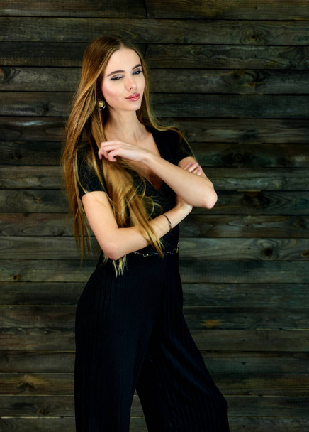 The concept of glamor, style. Model Stands in different poses in front of the camera. Vertical portrait of a pretty blonde girl with long hair and great makeup on a wooden background. - Photo, Image