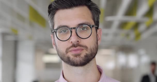 Close up of bearded caucasian man in 30s looking to camera and blinking. Portrait of handsome guy with serious face expression. Blurred modern office background. Concept of people. - Video