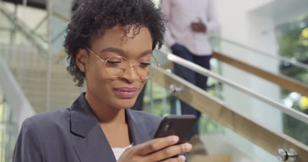 Close up of african businesswoman in glasses using phone and smiling in modern office building. Female office worker surfing net, swiping, texting at smartphone. Concept of people, tech. - Video, Çekim