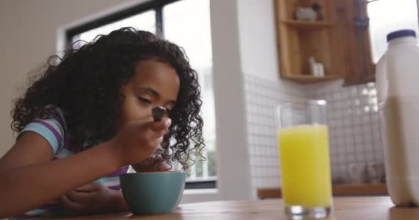 Side view close up of a young African American girl at home in the kitchen, sitting at the table eating a bowl of breakfast cereal, with a glass of orange juice on the table in front of her, slow motion - Filmagem, Vídeo