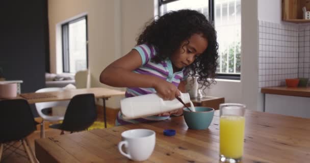 Side view of a young African American girl at home in the kitchen, sitting at a table pouring milk from a bottle onto her breakfast cereal and putting the lid back on the bottle, slow motion - Materiał filmowy, wideo