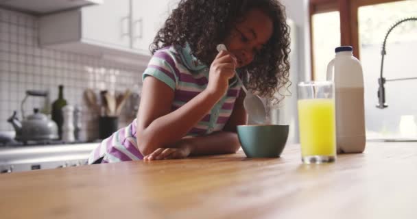 Side view of a young African American girl at home in the kitchen, sitting at the table with a bowl of breakfast cereal, mixing it with her spoon and smiling, a glass of orange juice on the table in front of her, slow motion - Filmati, video