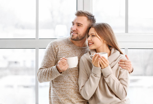 Ευτυχισμένο ζευγάρι με φλιτζάνια ζεστό τσάι αναπαύεται κοντά στο παράθυρο στο σπίτι - Φωτογραφία, εικόνα