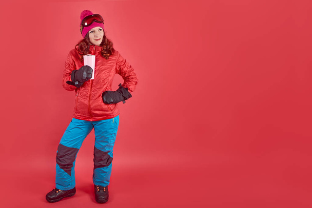 Side view studio shot portrait woman wearing rad ski clothes and mask standing in red background with tinsel - Zdjęcie, obraz