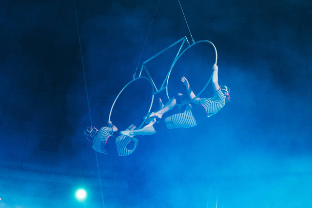 KYIV, UKRAINE - NOVEMBER 1, 2019: Low angle view of air gymnasts in smoke performing with rings in circus  - Photo, Image