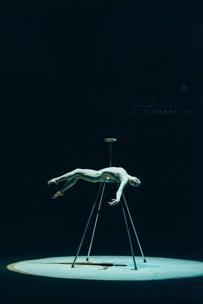 KYIV, UKRAINE - NOVEMBER 1, 2019: Side view of beautiful gymnast lying in spotlight in circus isolated on black - Foto, imagen
