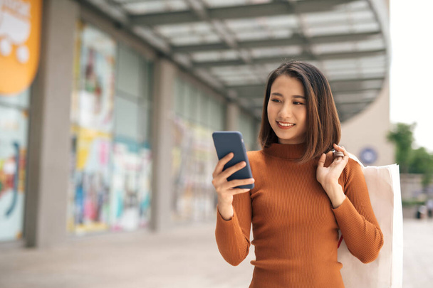 Портрет елегантної молодої азіатської жінки, яка тримає сумочки і користується смартфоном, залишаючи торговий центр. - Фото, зображення