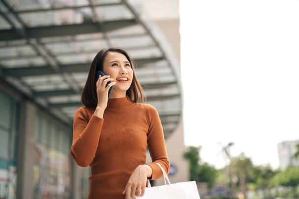 Portrait of happy young Asian woman talking on mobile and carrying paper shopping bags walking outdoor mall - Фото, зображення