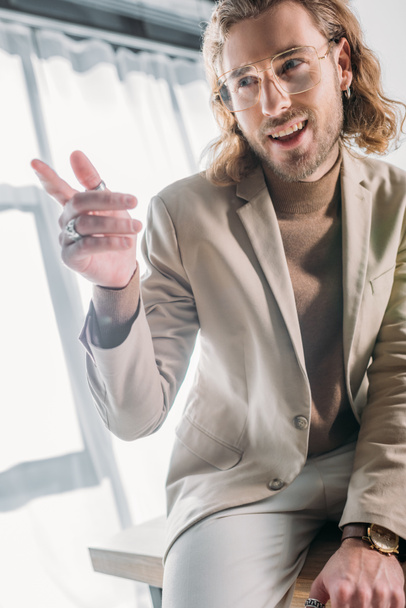 low angle view of smiling fashionable businessman sitting on desk and pointing with finger away in office - Photo, Image