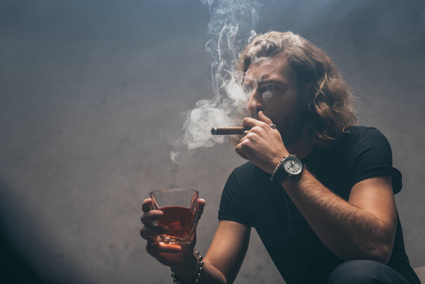 fashionable businessman in black outfit smoking cigar and drinking whiskey near grey wall - Photo, Image