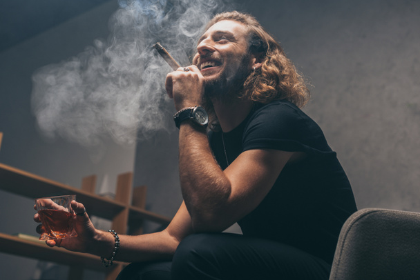low angle view of smiling fashionable businessman in black outfit smoking cigar and drinking whiskey - Fotó, kép