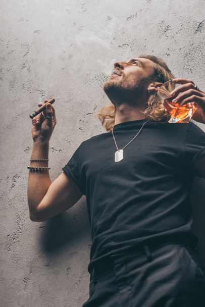 low angle view of fashionable businessman in black outfit smoking cigar and drinking whiskey near grey wall - Φωτογραφία, εικόνα