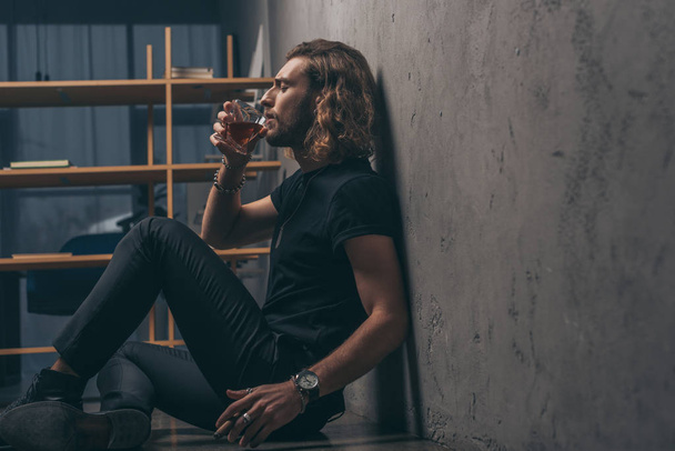side view of fashionable businessman in black outfit drinking whiskey on floor near grey wall - Fotografie, Obrázek