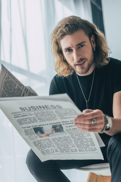 selective focus of smiling fashionable casual businessman in total black outfit holding business newspaper in office - Фото, изображение