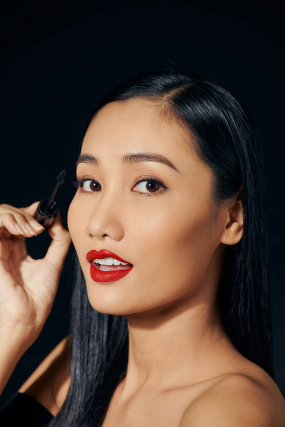 Közelkép portré szép aranyos aranyos édes vonzó vidám pozitív ázsiai nő gazdaság a kezében alkalmazó trendi fekete szempillaspirál elszigetelt - Fotó, kép
