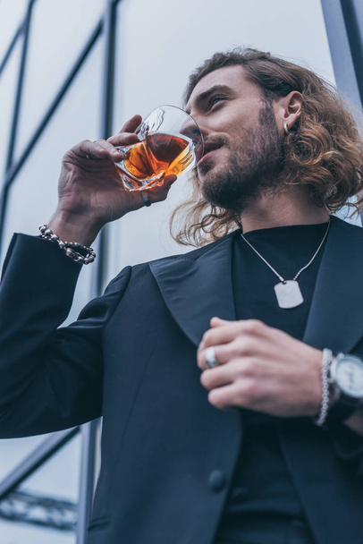 low angle view of fashionable businessman in black suit drinking whiskey near office building - Фото, зображення