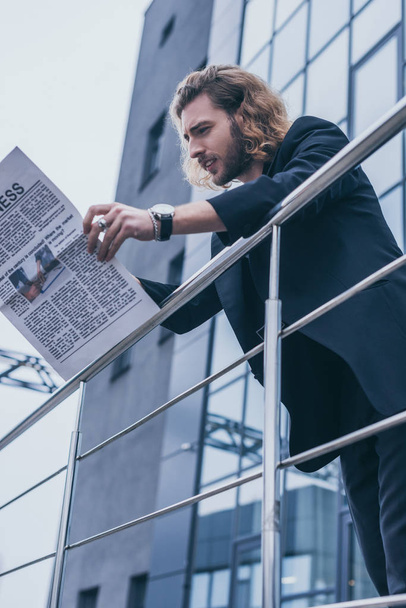 low angle view of fashionable businessman in black suit reading business newspaper near office building and railing - 写真・画像