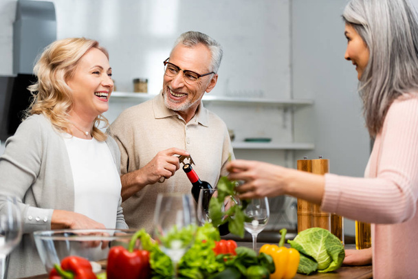 smiling woman cooking and talking with friend, man opening bottle with wine  - Photo, Image