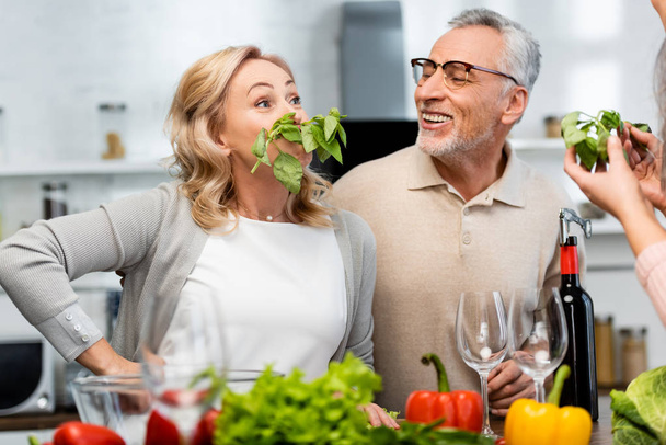 atractiva mujer con albahaca mirando a sus amigos sonrientes en la cocina
  - Foto, Imagen