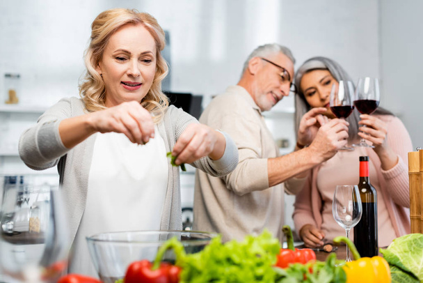 selective focus of woman adding lettuce to bowl and her multicultural friends looking at wine glasses on background  - Photo, Image