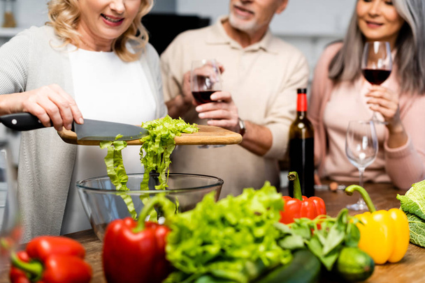 cropped view of woman adding lettuce to bowl and her friends holding wine glasses  - Фото, изображение