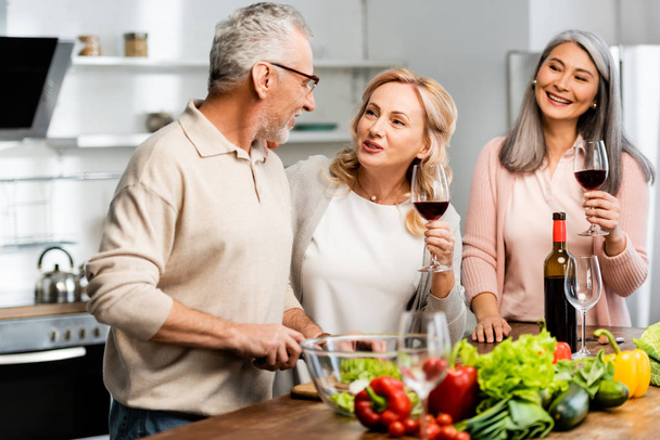 smiling multicultural woman holding wine glasses and man cutting lettuce in kitchen  - Photo, Image