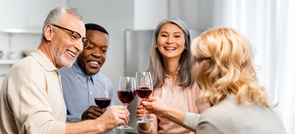Panoramaaufnahme lächelnder multikultureller Freunde, die mit Weingläsern in der Küche klirren  - Foto, Bild