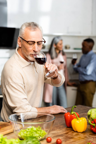 selective focus of man drinking wine and multicultural friends talking on background  - Photo, Image