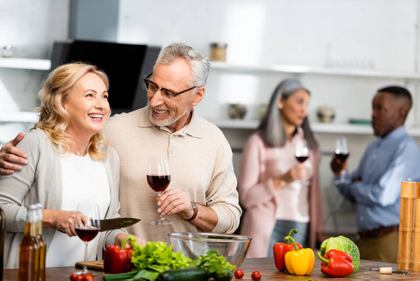 вибірковий фокус усміхненого чоловіка, який тримає келих для вина та жінку, тримає ніж, багатокультурні друзі говорять на фоні
  - Фото, зображення