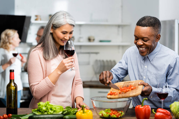 smiling african american man adding cut bell pepper to bowl and asian woman smelling wine  - Photo, Image
