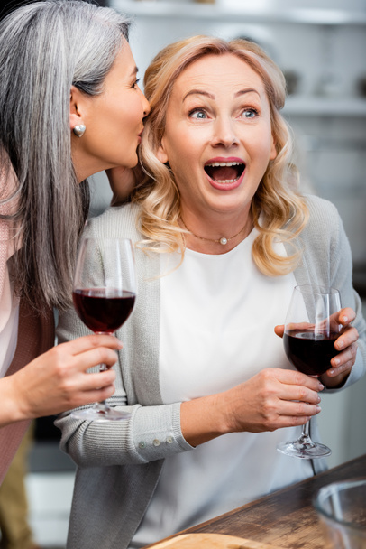 asian woman telling secret to shocked friend with wine glass  - Foto, Bild