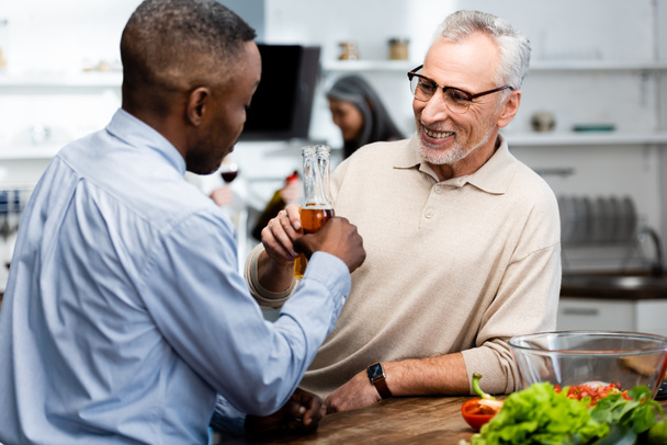 african american man clinking with his smiling friend in kitchen  - Foto, immagini