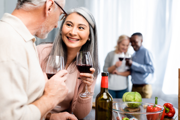 selective focus of smiling multicultural friends talking and holding wine glasses  - Foto, Bild