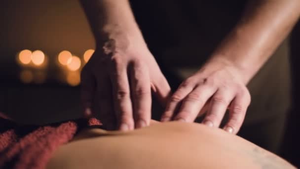 Young male massage therapist is doing finger massage of a woman back with a tattoo in a massage room with dim light on the background of candles. Low key premium massage concept - Filmagem, Vídeo