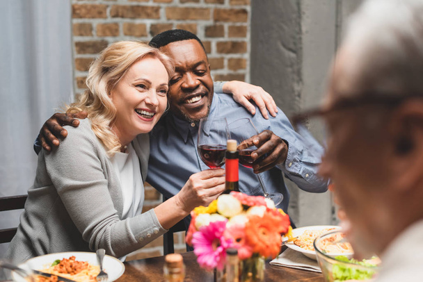 selektywne skupienie się na uśmiechniętych wielokulturowych przyjaciołach przytulających się i brzęczących podczas kolacji  - Zdjęcie, obraz