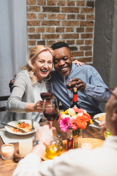 enfoque selectivo de amigos multiculturales sonrientes abrazos y tintineo durante la cena
  - Foto, Imagen