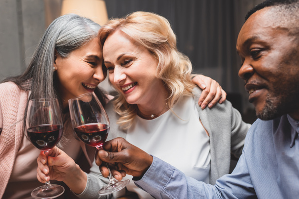 usmívající se multikulturní přátelé objímající a cinkající sklenicemi na víno během večeře  - Fotografie, Obrázek