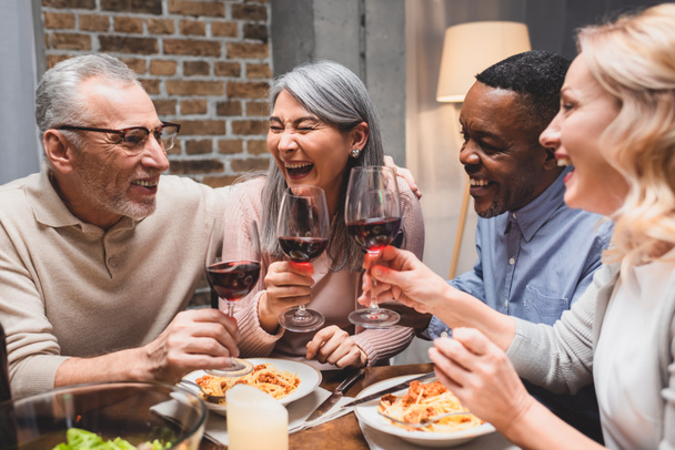 Lächelnde multikulturelle Freunde, die beim Abendessen mit Weingläsern plaudern und klirren  - Foto, Bild