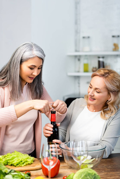 uśmiechnięci wielokulturowi przyjaciele otwierający butelkę wina korkociągiem w kuchni  - Zdjęcie, obraz