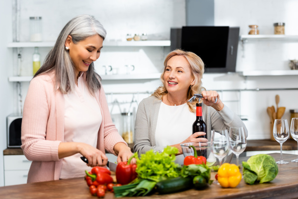 smiling woman opening wine bottle with corkscrew and looking at her asian friend with knife  - Photo, Image