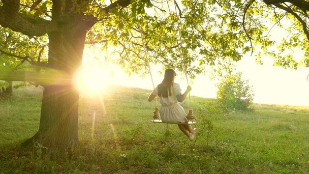 Genç kız güneşin altında meşe dalında sallanıyor. Uçma hayalleri. Mutlu çocukluk kavramı. Parkta beyaz elbiseli güzel bir kız. Genç kız yaz akşamı ormanda salıncakta sallanmaktan hoşlanıyor. - Fotoğraf, Görsel